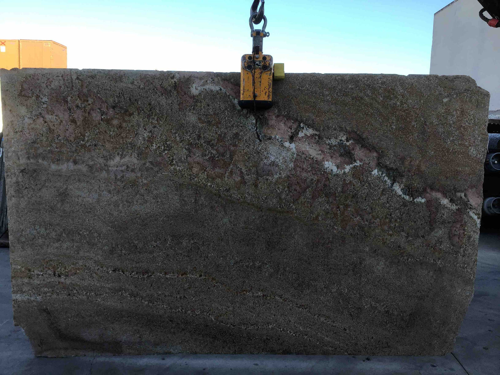 Granite slab Costa Dourada being lifted by a yellow clamp; rough brown stone with pink veining, 115 x 61 cm